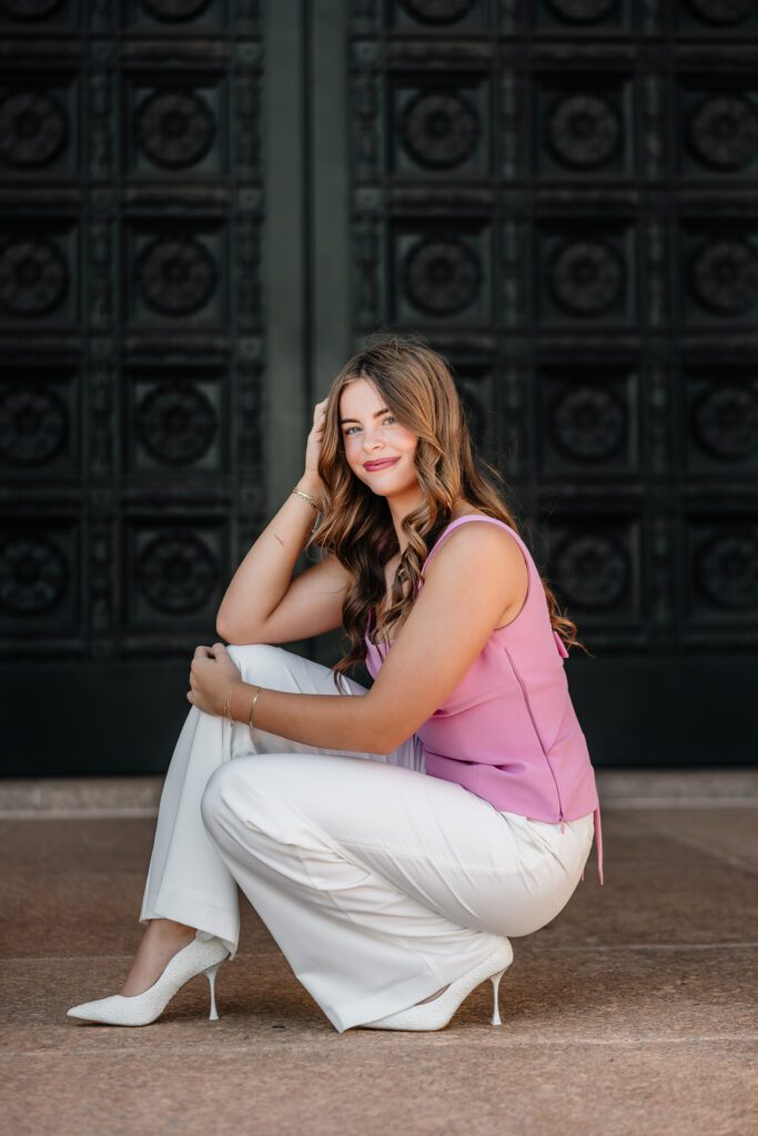 Senior girl in a pink top and white pants crouching against the carved dark doors of the Oklahoma State Capitol, OKC senior portrait by Ellie Piper