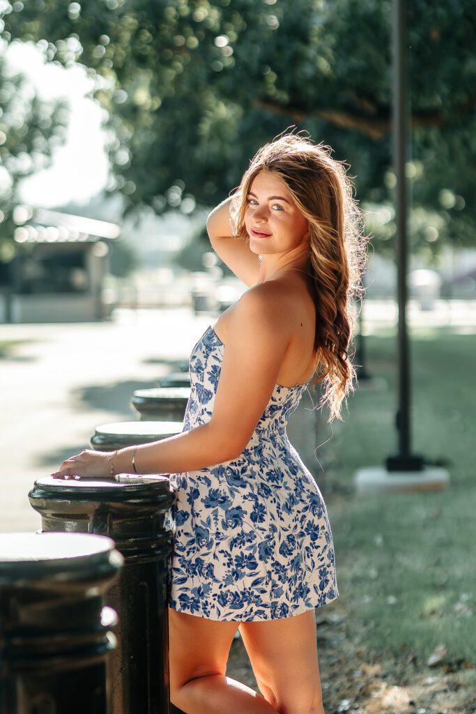 High school senior in a blue and white floral strapless dress looking over her shoulder beside black bollards at golden hour, senior portrait in Edmond by Oklahoma photographer Ellie Piper