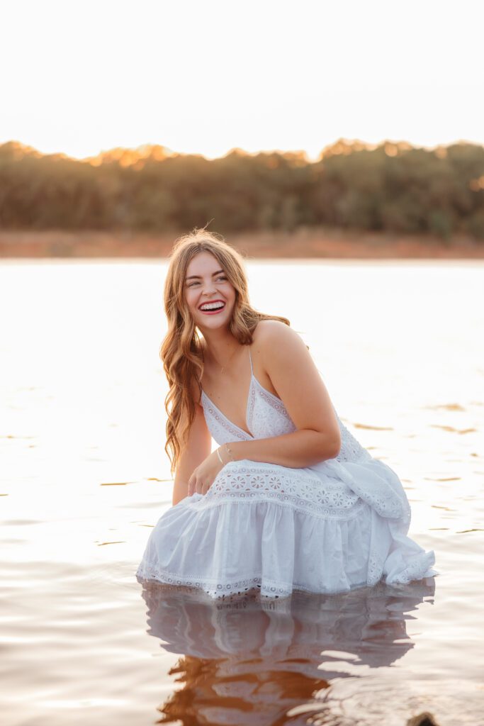 Laughing senior in a white eyelet dress kneeling in the water at Lake Arcadia at sunset, Edmond senior photos by Ellie Piper
